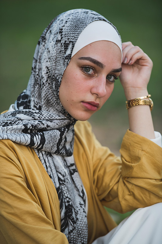 A hijabi girl staring at camera in her EMMA Scarf Black Python while resting her head on her hand in a yellow top