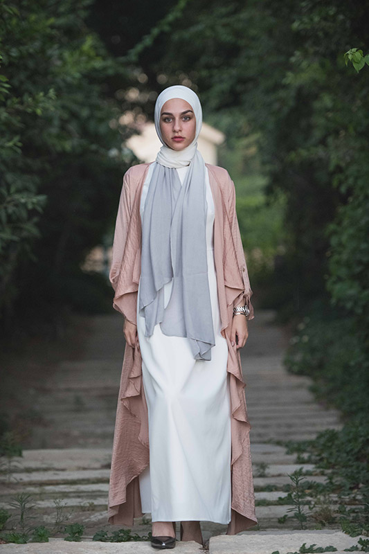 A hijabi girl in EMA Scarf Powdered Slate, a white maxi dress and a pink frill kimino in the forest staring at camera