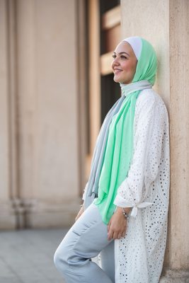 White Hijabi wearing EMMA Scarf Apple crunch and a white lace kimono, leaning against a walla wakk