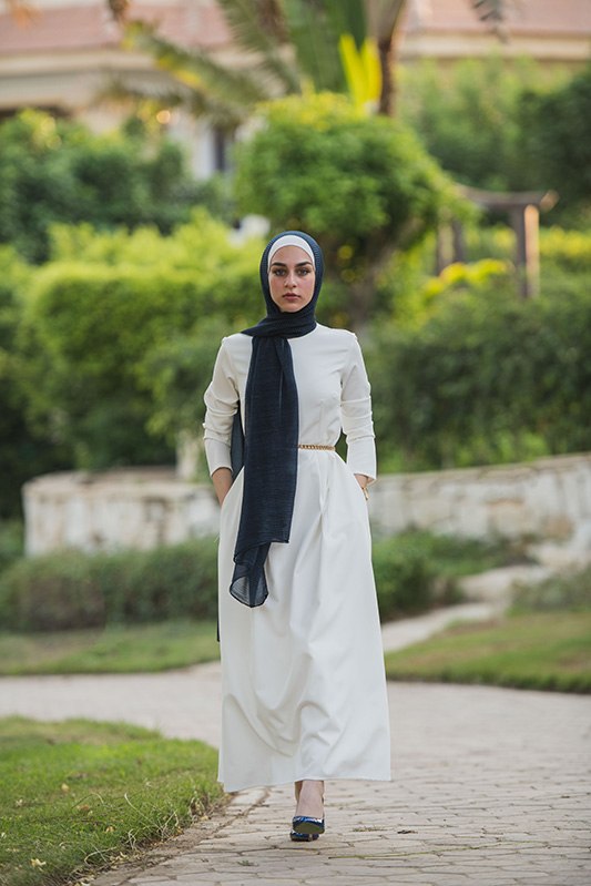 Hijabi Model in EMMA Scarf Navy Shimemr ,a white maxi dress , walking towards camera , hands in her pocket in a garden