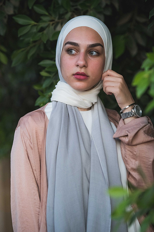 A close up of a white hijabi girl in EMMA scarf Powdered Slate fixing her hijab, staring at a distance