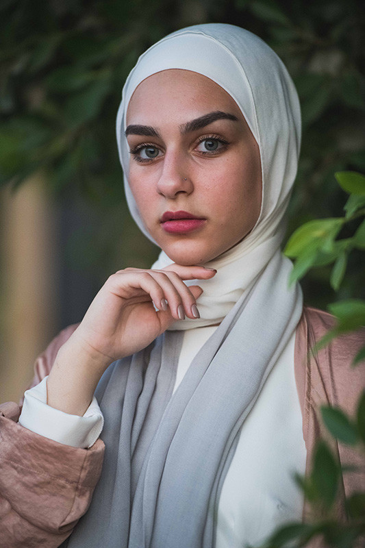 A close up of a white hijabi girl in EMMA scarf Powdered Slate with her pointer finger under her chin , staring at the camera